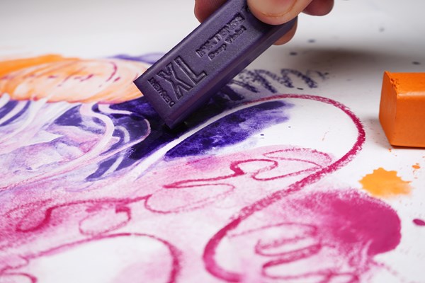 A hand uses a purple Derwent Inktense XL block to draw squiggles on a white surface.
